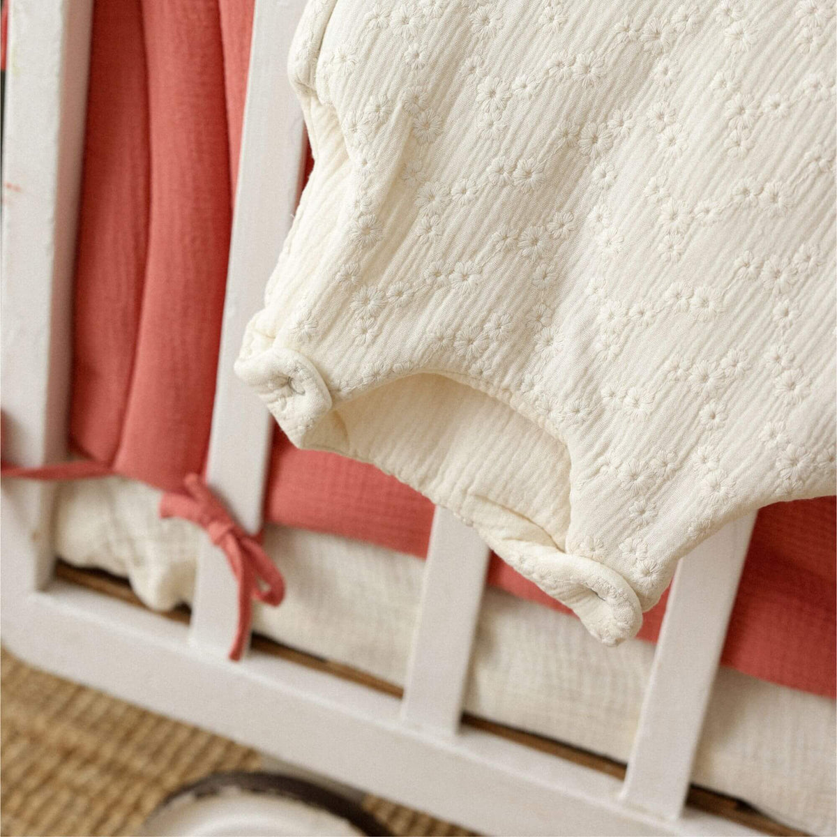 Détail d’une gigoteuse en gaze de coton brodée écrue, avec pressions sur le bas, posée sur un lit bébé blanc et un linge de lit en gaze terracotta