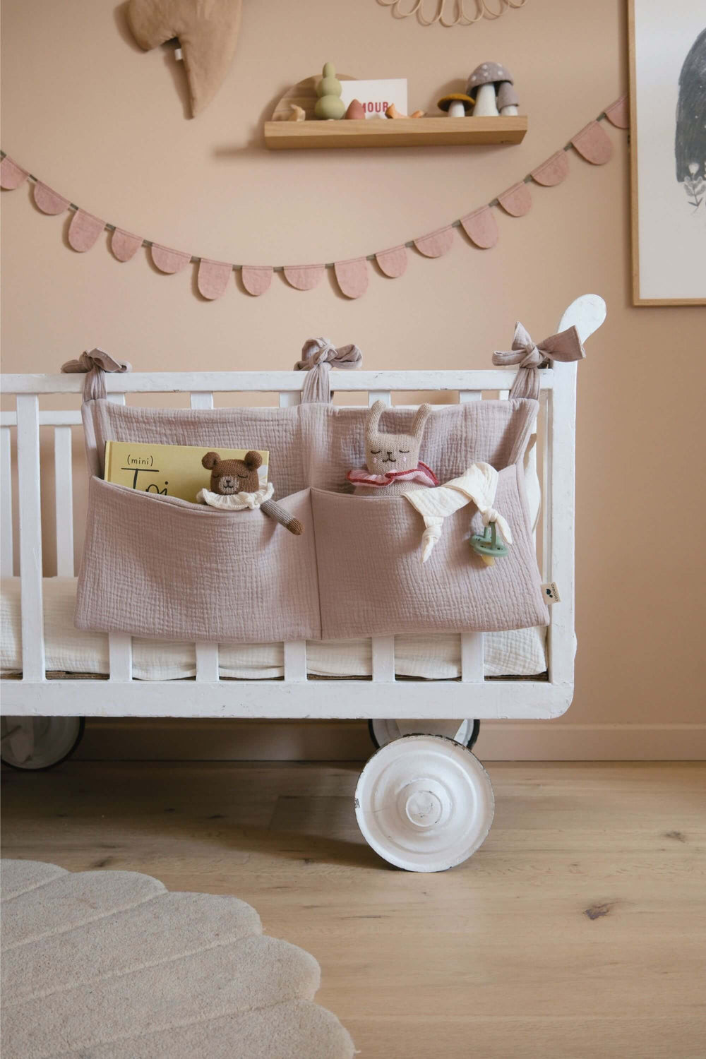 Range-doudou en gaze de coton rose poudré accroché à un lit de bébé, avec doudous et livre dans ses poches